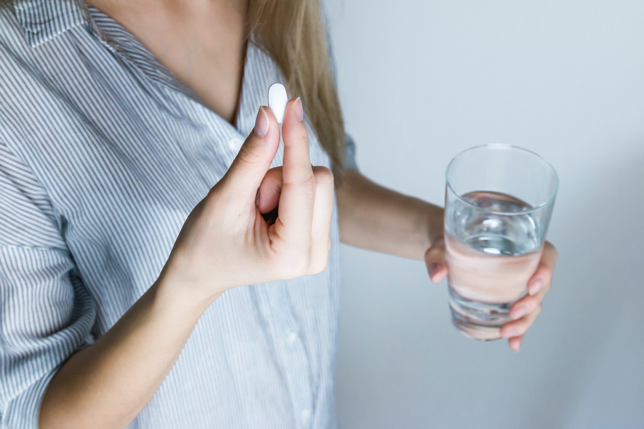 Young woman in a striped shirt holds a white pill and a glass of water