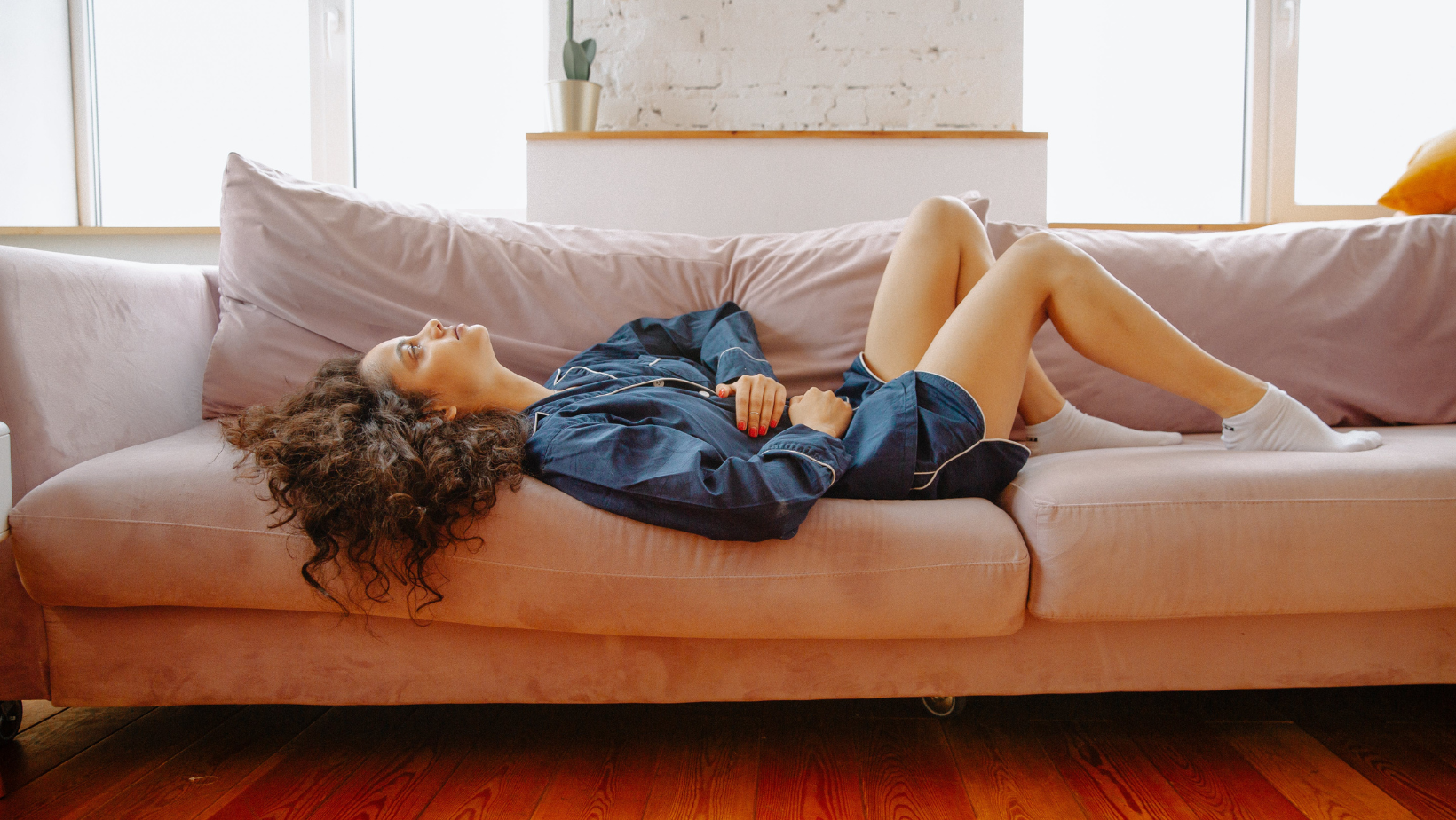 Young woman in blue pajamas lays on a couch holding her abdomen in discomfort 