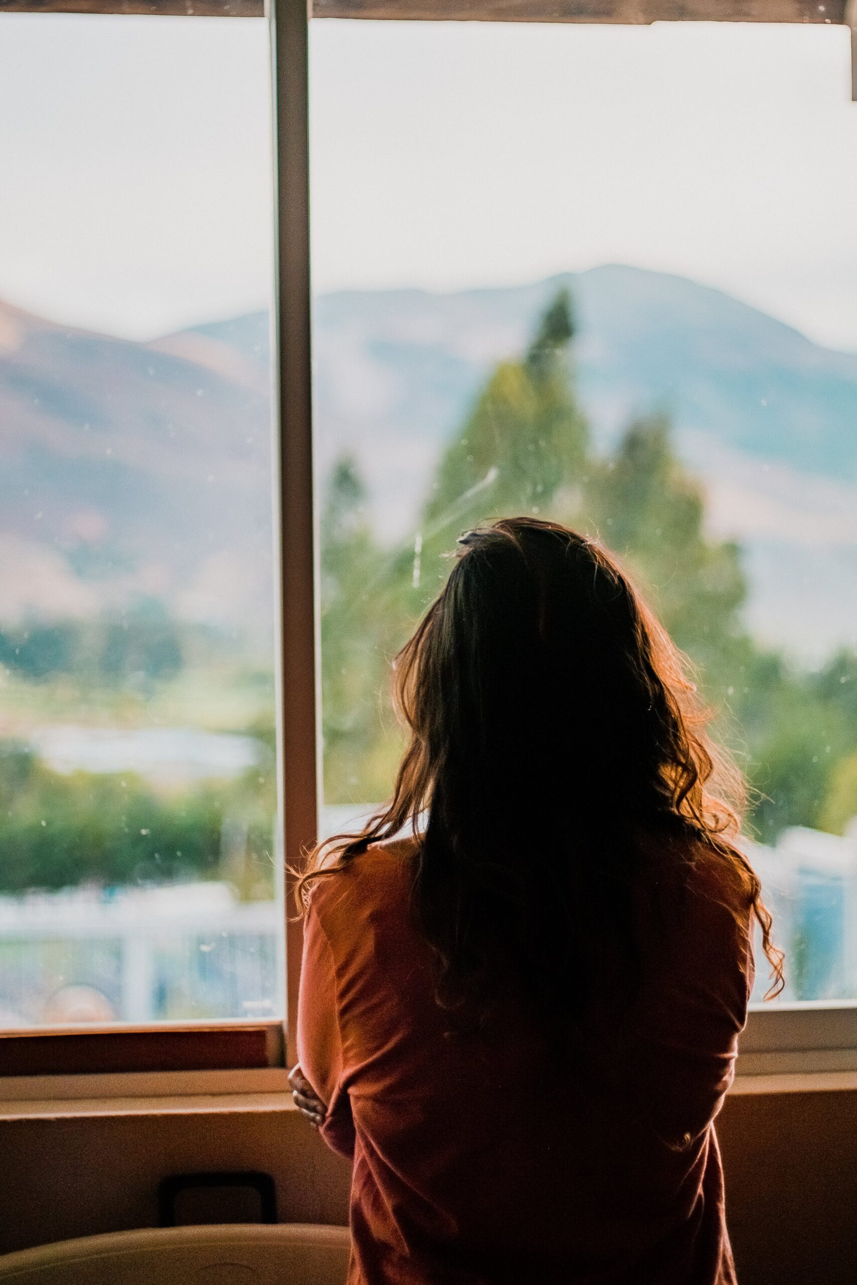 Young woman with long, dark hair looks out a window at an out-of-focus outdoor scene
