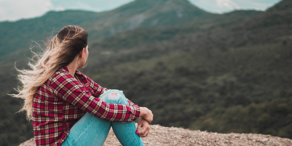 Young woman in a plaid shirt and torn jeans set on a boulder and looks out over a hilly, forested area in deep thought. 