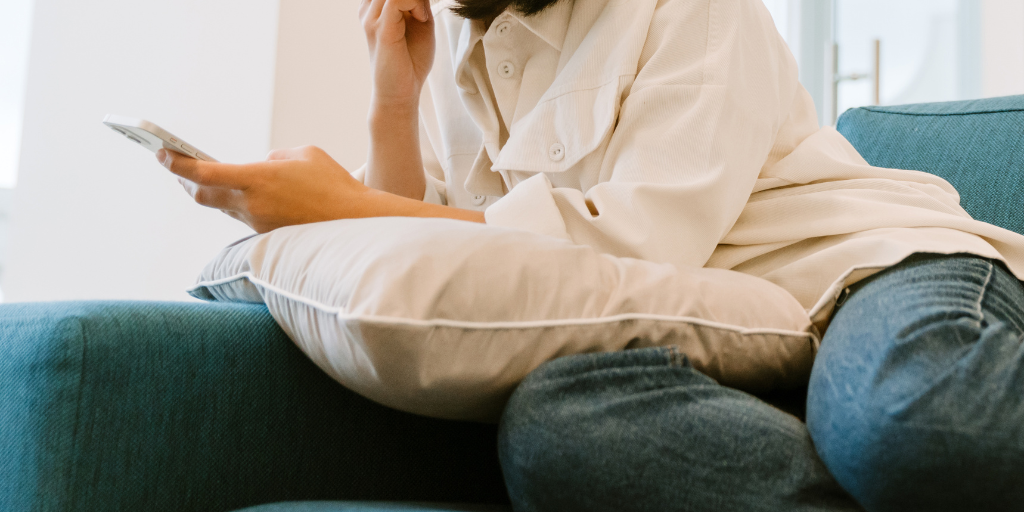 Woman in jeans and white shirt sets on teal couch holding a pillow and her phone