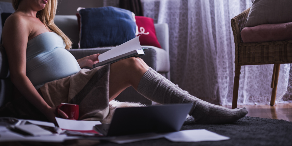 Pregnant student studies at home while setting on the floor surrounded by books, her computer, and a coffee mug