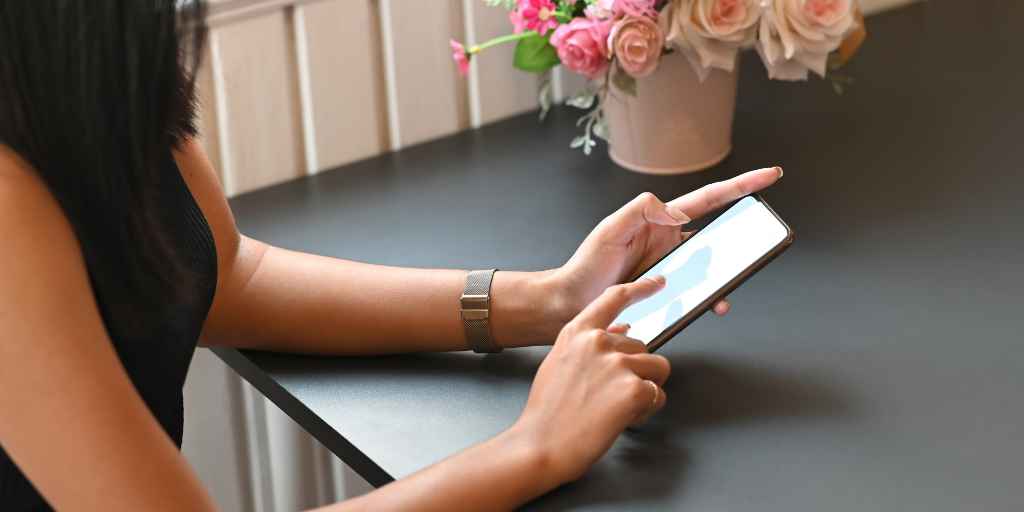 Woman searches her smart phone; pot of flowers on the table next to her